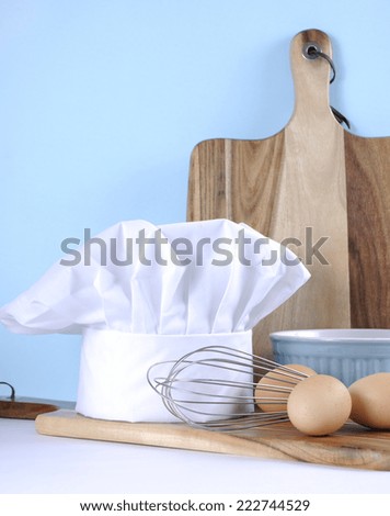 Modern kitchen cooking kitchenware and chef\'s hat with mixing bowl, whisk, chopping boards and eggs on a pale aqua blue and white background. Vertical.