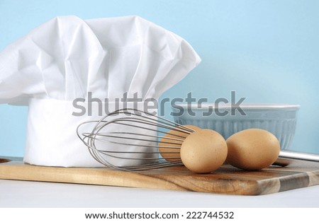 Modern kitchen cooking kitchenware and chef\'s hat with mixing bowl, whisk, chopping boards and eggs on a pale aqua blue and white background. Close up with copy space.