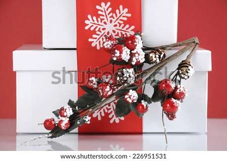 Stack of festive red and white theme Christmas gift boxes on reflective white table against a red background. Closeup on holly decoration.