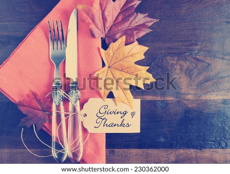 Retro vintage style Thanksgiving table place setting with greeting tag, orange napkin, autumn fall leaves on rustic dark recycled wood background, with copy space.