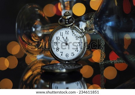 Happy New Year Eve party with pocket watch with five to midnight time and martini cocktail glasses on reflective table against bright color festive bokeh lights. Closeup.