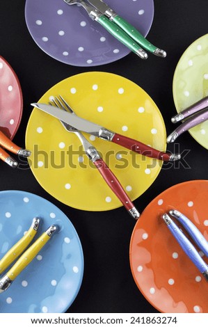 Dinner party table setting with red, blue, yellow, orange, green and purple bright polka dot plates and cutlery on black table cloth. Vertical.