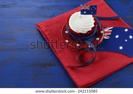 Happy Australia Day, January 26, theme table setting with red, white and blue cupcake on red polka dot plate and Australian flag decoration on dark blue wood background.