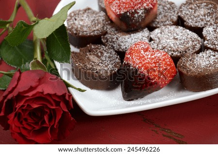 Happy Valentines Day chocolate dipped heart shaped strawberries with chocolate roulade swiss roll on heart shape plate and red rose bud on red vintage wood background, closeup.