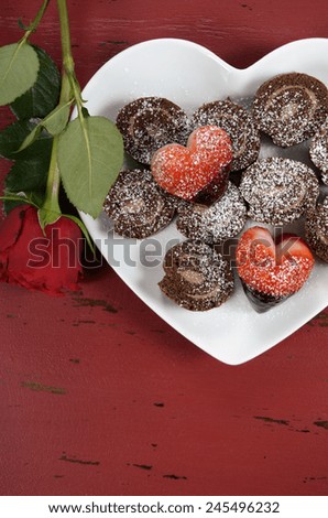 Happy Valentines Day chocolate dipped heart shaped strawberries with chocolate roulade swiss roll on heart shape plate and red rose bud on red vintage wood background, vertical overhead.