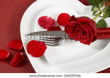 Romantic Valentines Day table place setting with white heart shape plates with rose and petals on red wood background.