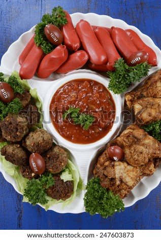 Super Bowl Sunday football party celebration food platter with chicken buffalo wings, meat balls, hot dogs and salsa dip on blue wood table, vertical overhead.