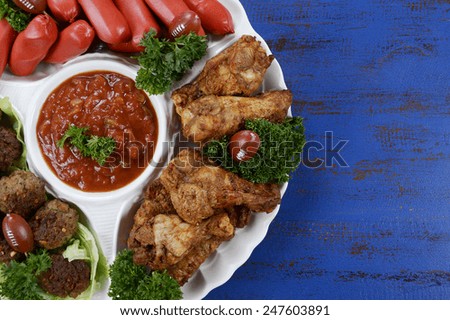 Super Bowl Sunday football party celebration food platter with chicken buffalo wings, meat balls, hot dogs and salsa dip on blue wood table.