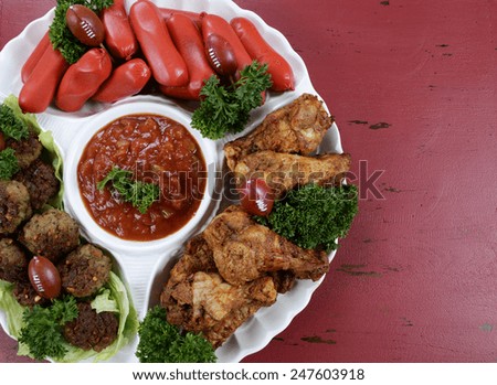 Super Bowl Sunday football party celebration food platter with chicken buffalo wings, meat balls, hot dogs and salsa dip on red wood table, with copy space.