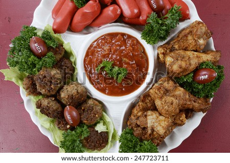 Super Bowl Sunday football party celebration food platter with chicken buffalo wings, meat balls, hot dogs and salsa dip on red wood table, overhead.