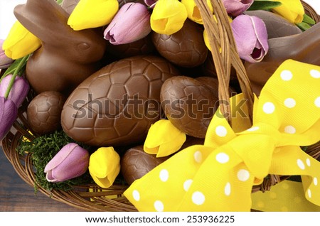 Happy Easter chocolate hamper of eggs and bunny rabbits in large basket with yellow and pink purple silk tulip flowers on dark wood table, overhead.