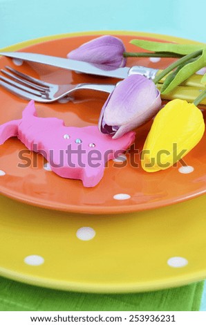 Happy Easter bright color orange, yellow and green polka dot theme table place setting with pink fondant bunny cookie and tulips on pale aqua blue distressed wood table, vertical closeup.