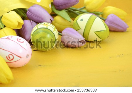 Happy Easter background with painted Easter eggs, yellow and purple silk tulips and polka dot ribbon on vintage style rustic distressed yellow wood table, with copy space for your text here, closeup.