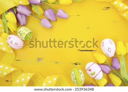 Happy Easter background with painted Easter eggs, yellow and purple silk tulips and polka dot ribbon on vintage style rustic distressed yellow wood table, with copy space for your text here.
