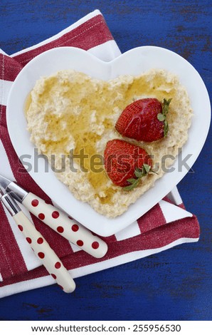 Plate of nutritious and healthy cooked breakfast oats with strawberries and honey in heart shaped bowl on dark blue rustic wood table, vertical.