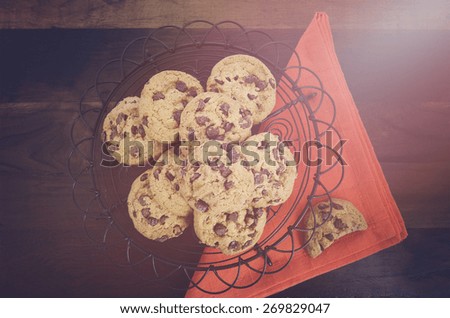 Chocolate chip cookies on vintage baking rack with orange napkin on dark wood background, with applied vintage style filters and added light stream.