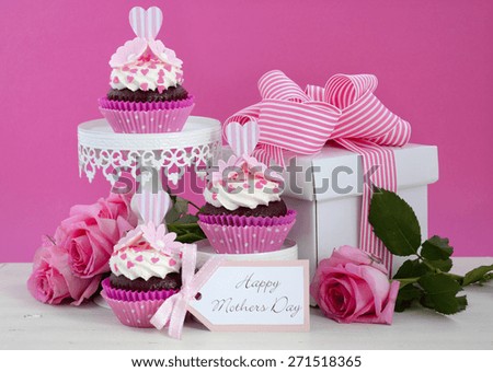 Happy Mothers Day pink and white cupcakes on retro style cake stands and large gift box on vintage white wood table.