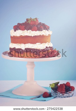 Fresh whipped cream and berries layer sponge cake on pink cake stand against pale blue and white background, with applied retro style filters and added lens flare light beam.
