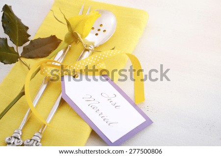 Yellow and white theme wedding table place setting with antique silverware, napkin and yellow rose bud on white shabby chic table, with applied retro style filters and added lens flare.