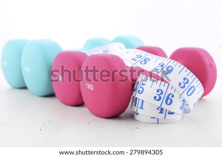 Health and fitness concept with pink and blue dumbbells and tape measure on white distressed wood table background.