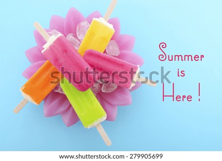 Summer is Here concept with bright color ice pop, ice creams on ice in pretty pink bowl blue background with text.