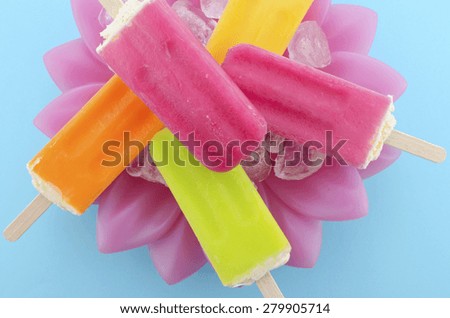 Summer is Here concept with bright color ice pop, ice creams on ice in pretty pink bowl blue background, overhead view.close up.