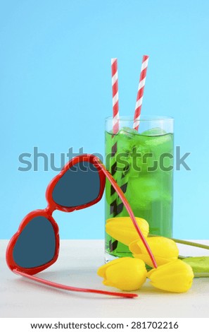 Summer Is Here concept with red heart shape sunglasses and yellow flowers and tall cool drink on white wood table and sky blue background.