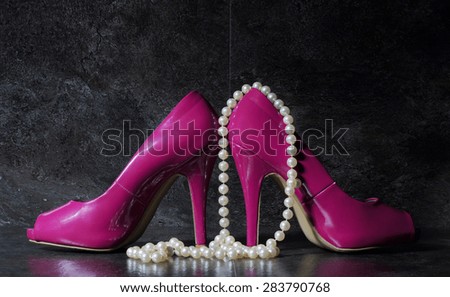 Glamorous pair of ladies pink high heels with long strand of white pears against a dramatic black slate background still life.