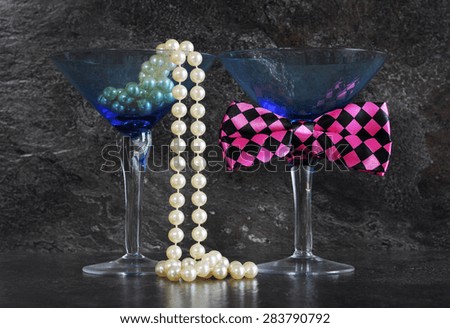 Ladies and Gentleman\'s vintage blue martini glasses with pearls and fun pink check bow tie  on black slate background.