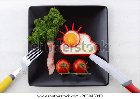 Bright happy breakfast with sunny egg, bacon tree with parsley leaves and cherry tomato flowers on black square plate on white wood table.