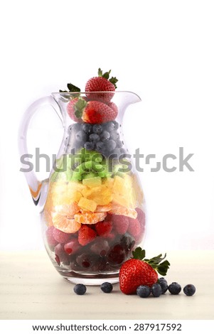 Fresh fruit juice concept with rainbow color fruit including cherries, strawberries, raspberries, mandarin segments, kiwi fruit, and blueberries in tall glass jug, on white shabby chic wood table.