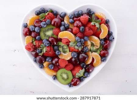 Colorful rainbow fruit including raspberries, strawberries, cherries, blueberries, mandarins and kiwi fruit  in heart shape bowl on white shabby chic distressed table, overhead view.