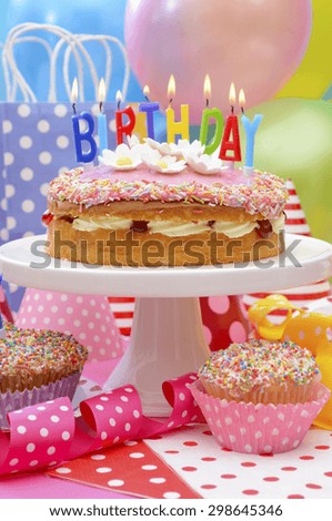 Bright colorful Happy Birthday Party Table with balloons, streamers, party favor gift bags and birthday cake with lit candles.