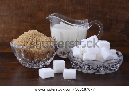 Cream and sugar in crystal glassware with coffee sugar crystals and sugar cubes on dark vintage wood background.