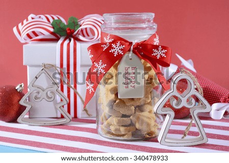 Christmas gift of a glass jar of star shaped shortbread cookies with happy holidays gift tag and ornaments on red, white and pale blue background with bright side lighting shining through jar.