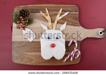 Christmas fun for the children with reindeer face cut out of bread sandwich with decorations on chopping board on a rustic red wood table.