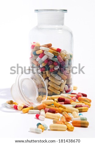 Variety of pharmaceutical capsules and pills lying on a white surface with an open half filled glass jar behind in a health care concept