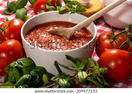 Bowl of fresh healthy homemade tomato puree with a wooden spoon for use in Italian cuisine surrounded by fresh basil for seasoning and ripe red tomatoes