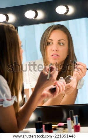 Attractive girl with long brunette hair applying her makeup in a mirror admiring her reflection in a beauty concept, view from behind of the reflection
