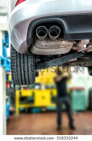 Mechanic repairing a motor car on a hoist in a workshop with focus to the rear exhaust pipe with the mechanic working on the front of the vehicle