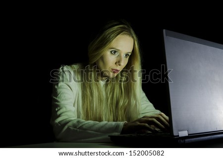 woman working on a computer by night in a dark room with only light from computer falling on her face horizontally cropped