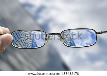 Out Of Focus Glass Business Building With Sky And Clouds Above And Hand Holding Glasses That Correct The Vision