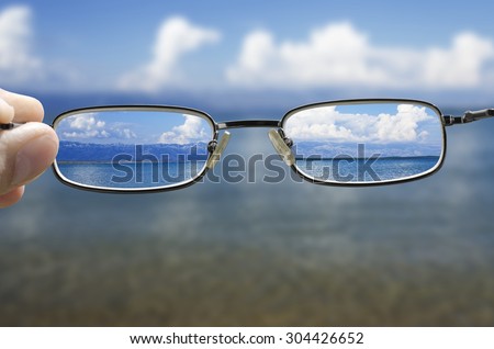blurry vision of a seashore on a beautiful summer day and a hand holding a pair of glasses that correct the vision