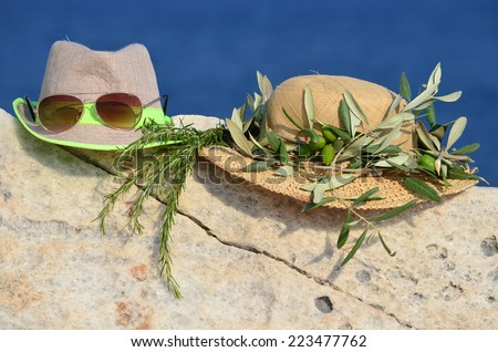 Summer vacation concept, two hats, sunglasses, olive wreath and sprig of rosemary on marble rock against blue bokeh background
