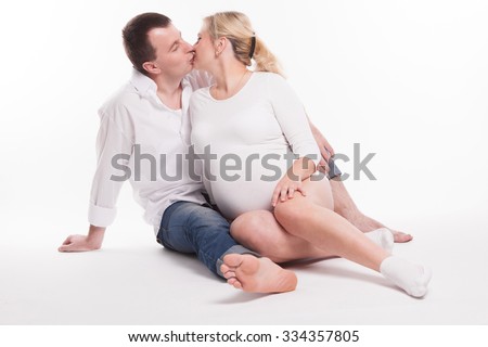 young dad in jeans and a white shirt and a pregnant woman in a white shirt sitting and gently kiss on a white background