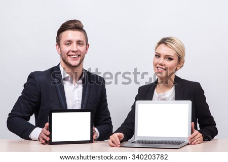 a man in a business suit sitting at a desk and holding a a tablet, sitting next to a woman in a business suit and blonde demonstrates laptop screen