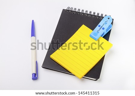 closed black cover notebook ,yellow empty post note, pen and blue clip with white background
