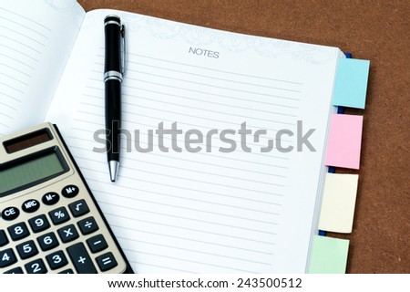 Blank notebook page with black ball pen, calculator and multi colored post it on wooden background