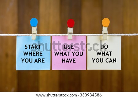Word quotes of START WHERE YOU ARE, USE WHAT YOU HAVE, DO WHAT YOU CAN on colorful sticky papers hanging by a rope against blurred wooden background.
