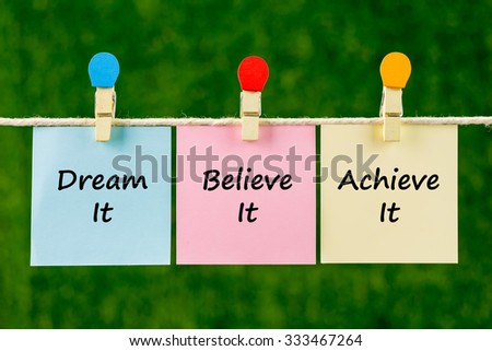 Word quotes of Dream It, Believe It, Achieve It on sticky color papers hanging on rope against blurred green background.